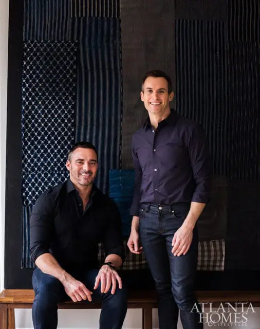 Two men smiling in front of a textile wall hanging.