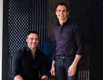 Two men smiling in front of a backdrop of patterned textiles.