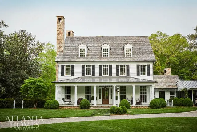 White farmhouse with stone chimney, wraparound porch, and manicured lawn.