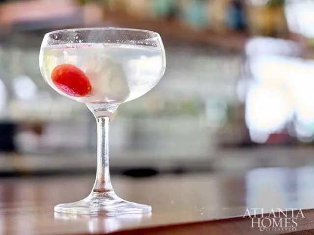 Clear cocktail with red cherry and ice in a coupe glass on a wooden bar
