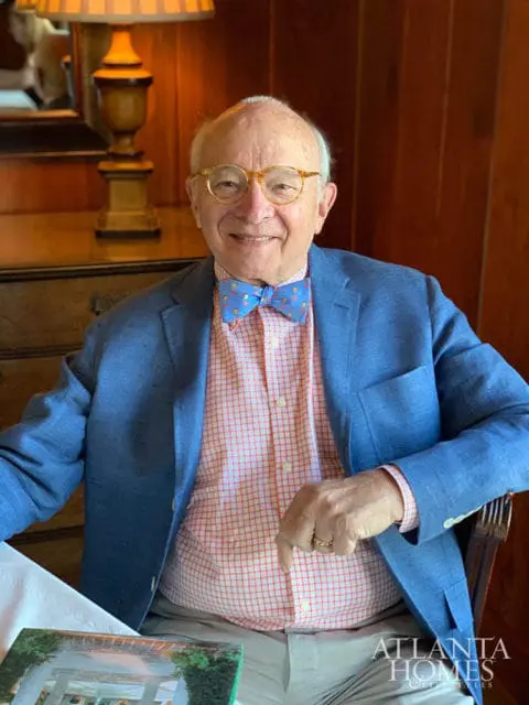 Elderly man with glasses and a bow tie smiles, wearing a blue blazer over an orange checkered shirt.