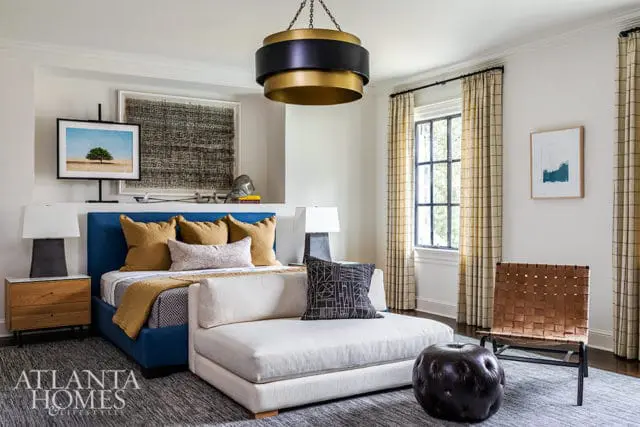 Blue upholstered bed with gold and patterned pillows, white sectional sofa, leather ottoman, and