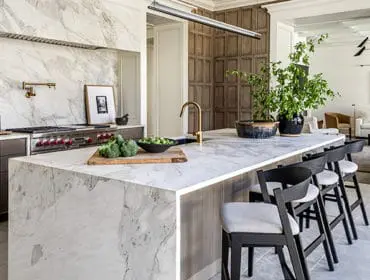 Marble island kitchen with stainless steel range, brass faucet, and dark wood bar stools.