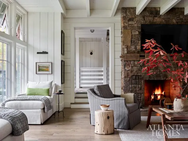 Living room with stone fireplace, white shiplap walls, and large windows.