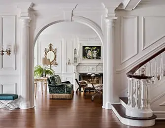 White foyer with arched doorway, fluted columns, dark wood floor, and velvet armchair.