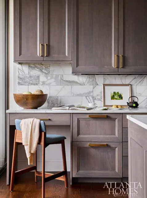 Gray kitchen cabinets with brass hardware, marble backsplash, and a wooden bowl of fruit.