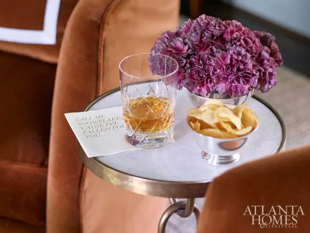 Side table with a glass of amber liquid, bowl of chips, purple flowers, and a cocktail napkin with