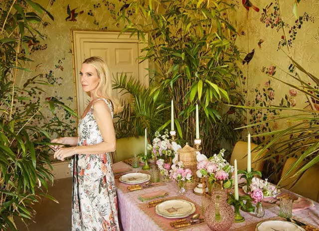 Woman in floral dress standing near a dining table set with pink linens and floral arrangements.