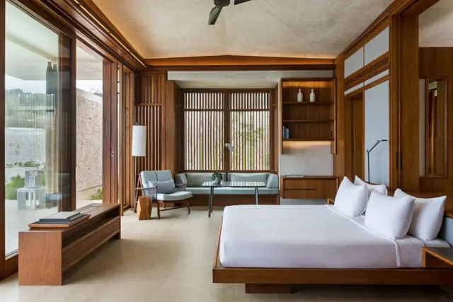 Bedroom with wood paneling, a white bed, light blue seating, and large windows.