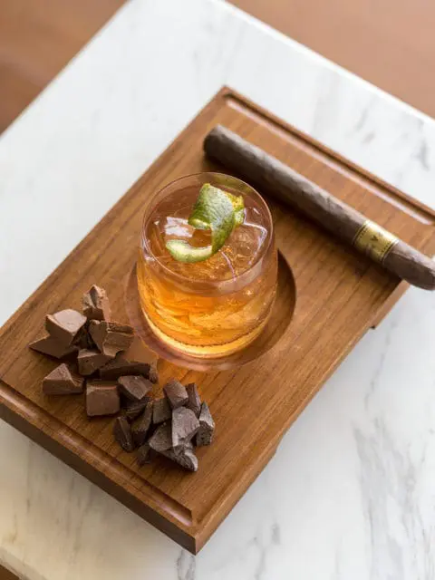 Whiskey cocktail with lime peel, chocolate chunks, and cigar on wooden tray