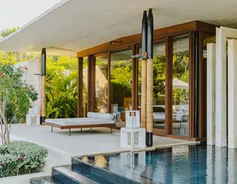 Modern living area with chaise lounge, white stools, and floor-to-ceiling windows overlooking lush