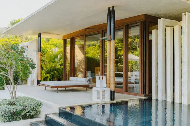 Modern house with concrete roof, wood-framed windows, and pool. Features a daybed and lush greenery.