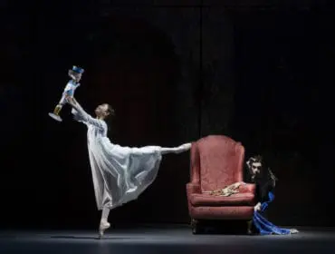 Ballerina in white dress holds a doll next to a pink armchair.