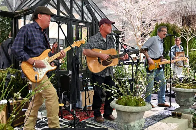 Band playing guitars and singing in a greenhouse with potted plants.