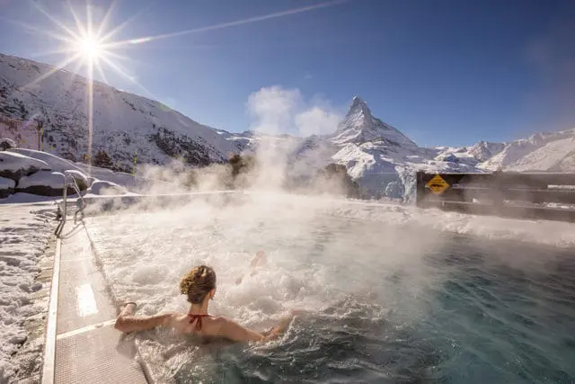 People in outdoor hot tub with steam rising, snow-covered mountains and Matterhorn peak in