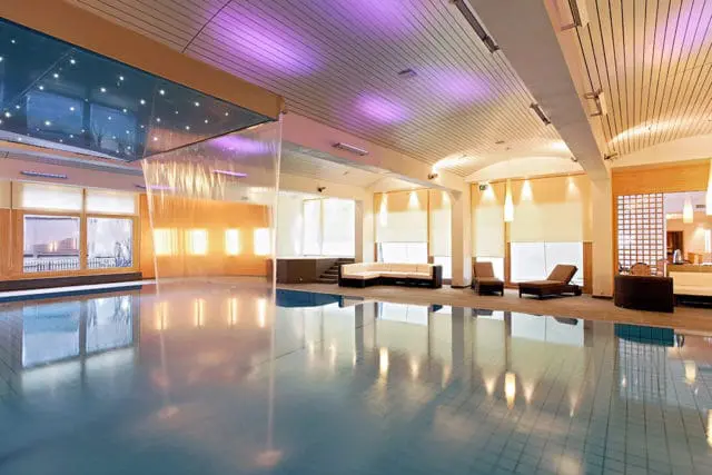 Indoor swimming pool with lounge chairs, sofas, and a waterfall feature. Purple lighting accents