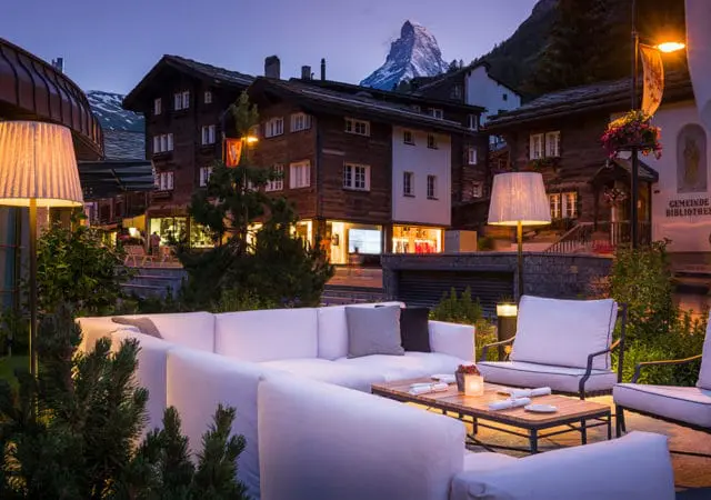 Outdoor seating area with white sofas and chairs, wooden buildings, and Matterhorn mountain at dusk.