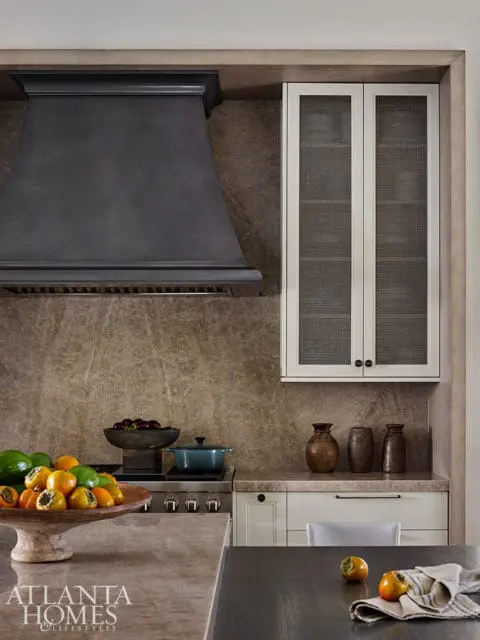 Kitchen with dark metal range hood, marble backsplash, and white cabinets with mesh fronts.