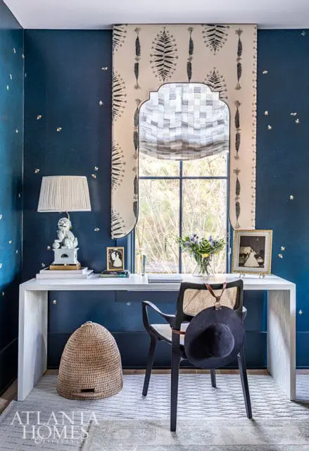 Blue desk area with patterned curtains, white desk, black chair with hat, and woven basket.