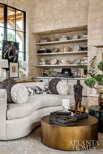 Living room with stone fireplace, curved sofa, brass coffee table, and shelves with decor.