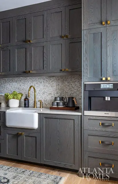 Gray wood kitchen cabinets with brass hardware, white farmhouse sink, and mosaic tile backsplash.