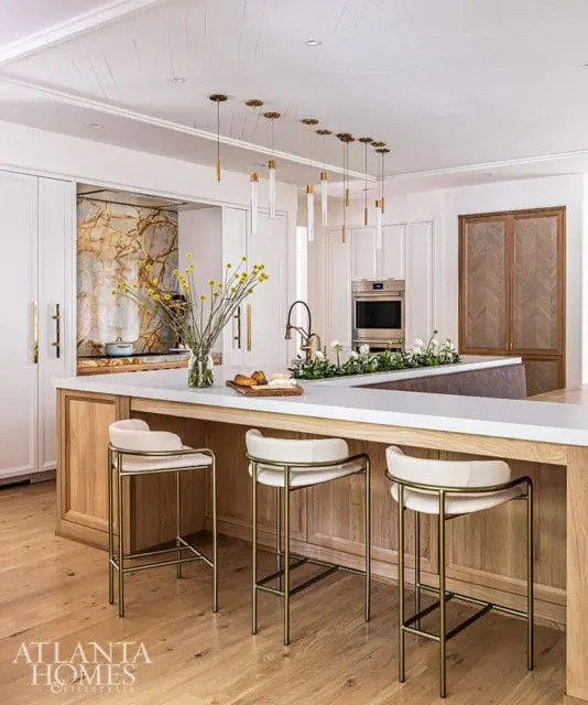 White kitchen island with wood cabinetry, white stools, and marble backsplash.