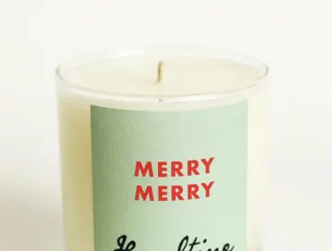 Glass candle with light yellow wax and a pale green label that reads 'MERRY MERRY Hazeltine' in red