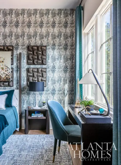 Bedroom with blue patterned wallpaper, teal velvet chair, and dark wood desk.