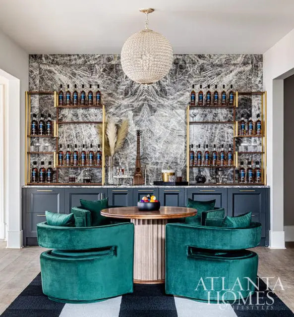 Bar area with dark cabinets, gold shelving, bottles, and green velvet chairs
