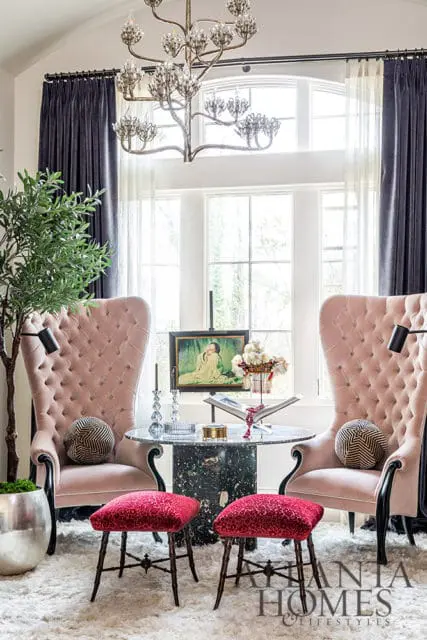 Two pink tufted wingback chairs flank a marble table with a painting and flowers.