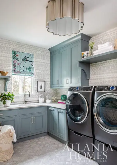 Laundry room with blue cabinets, gray washer and dryer, patterned wallpaper, and floral roman shade.