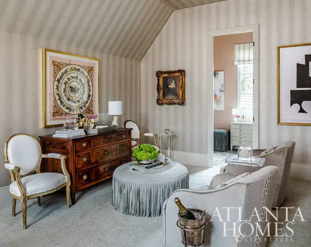 Living room with striped wallpaper, antique dresser, fringed ottoman, and plush chairs.