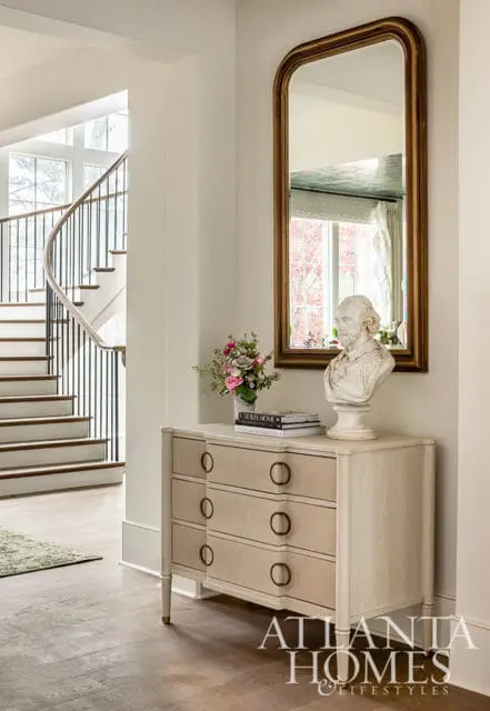 Hallway with curved staircase, light wood dresser, bust, and gold mirror
