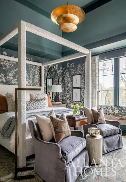 Bedroom with dark floral wallpaper, white canopy bed, gray velvet chairs, and brass chandelier.