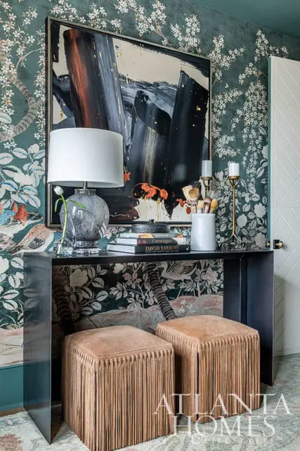 Entryway with dark floral wallpaper, abstract art, glass lamp, and two fringe ottomans.