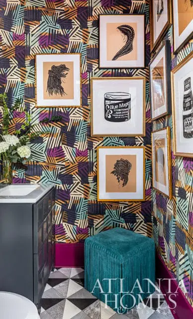 Bathroom with bold geometric wallpaper, framed art of Black hairstyles, and a teal fringed ottoman.