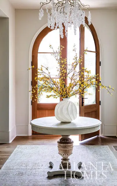 Entryway with arched wooden door, round table with white vase of yellow flowers, and crystal