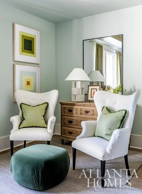 Two white wingback chairs with green pillows flank a wooden dresser with lamps.