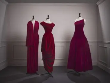 Three mannequins display red and magenta velvet gowns in a room with paneled walls.