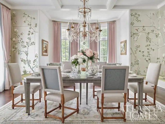 Dining room with floral wallpaper, pink curtains, and upholstered chairs around a table.