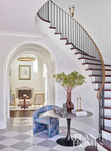 Interior foyer with curved staircase, blue patterned chair, and round table with vase and books.