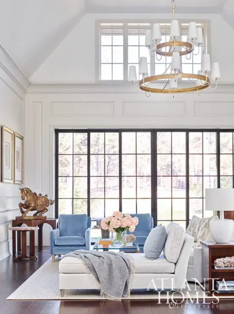 Living room with blue armchairs, white chaise, and large windows overlooking trees. Gold horse