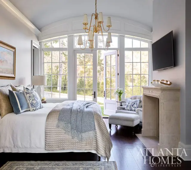 Bedroom with large windows, fireplace, tufted armchair, and a bed with blue and white pillows.