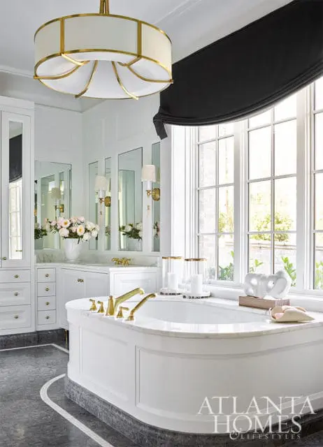 White bathroom with freestanding tub, marble countertops, brass fixtures, and large windows.