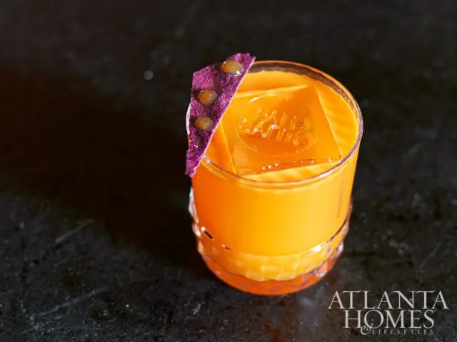 Orange cocktail with ice cube and purple garnish, Atlanta Homes & Lifestyles logo
