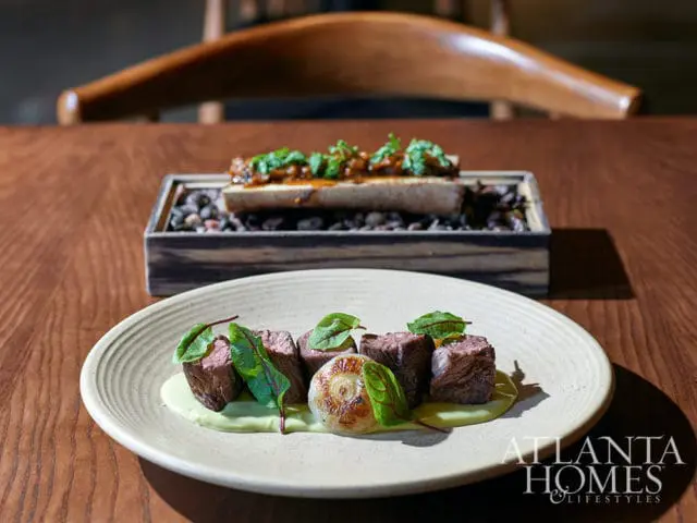Beef cubes with green puree and greens on a plate, with bone marrow in background.