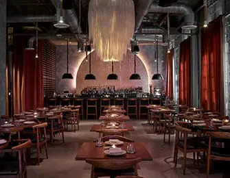 Restaurant dining room with wooden tables and chairs, bar in background, and large pendant light