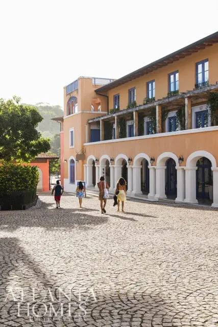 Orange building with arched colonnade and blue windows, people walking on cobblestone path.