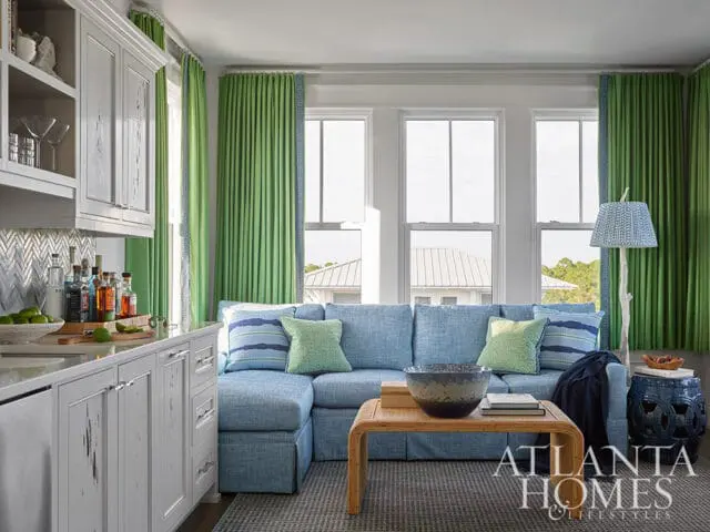 Blue sectional sofa with green and blue pillows, bamboo coffee table, and green curtains.