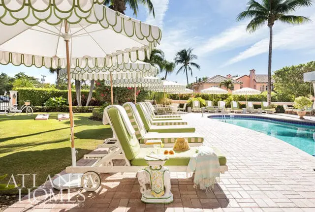 Lounge chairs and umbrellas by a pool with palm trees and a house in the background.